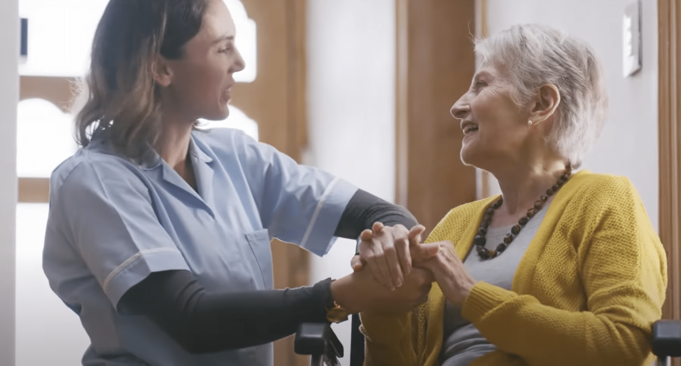 A Medical Assistant wearing a blue uniform smiles at an elderly woman in a wheelchair. The elderly woman, wearing a yellow cardigan, smiles back while holding the Medical Assistant's hands. They are in a well-lit room.